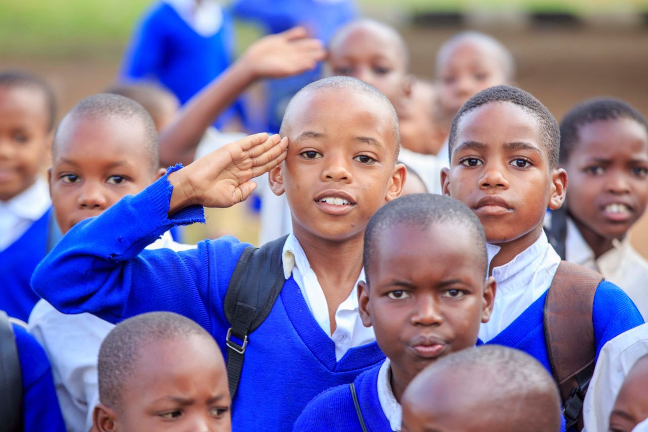 heros-img A group of school children in blue uniforms saluting outdoors on a sunny day.